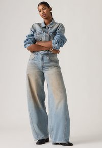 Νεαρή γυναίκα με πλεγμένα μαλλιά στέκεται με αυτοπεποίθηση φορώντας ξεθωριασμένο τζιν μπουφάν και τζιν παντελόνι με φαρδιά πόδια και μαύρα μπότες έως τον αστράγαλο.