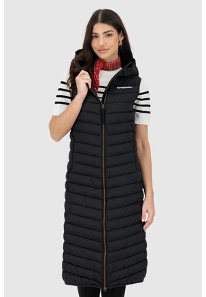 Frau trägt lange, schwarze, ärmellose Steppweste mit Kapuze über weißem Hemd mit schwarzen Streifen, hält Reißverschluss nahe am Kragen, roter Schal um den Hals.