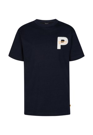 Blond jongetje staat, draagt een donkerblauw t-shirt met een witte letter P, beige korte broek, witte sokken met strepen en witte sneakers.