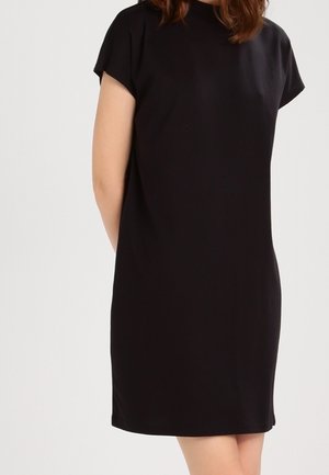 Woman wearing a short-sleeve, knee-length black dress standing against a plain white background.