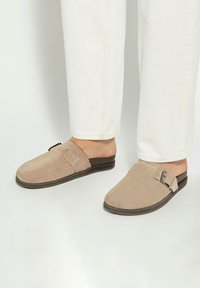 Beige suede slip-on shoes with a flat sole, featuring a side buckle detail and a smooth upper, paired with white pants.