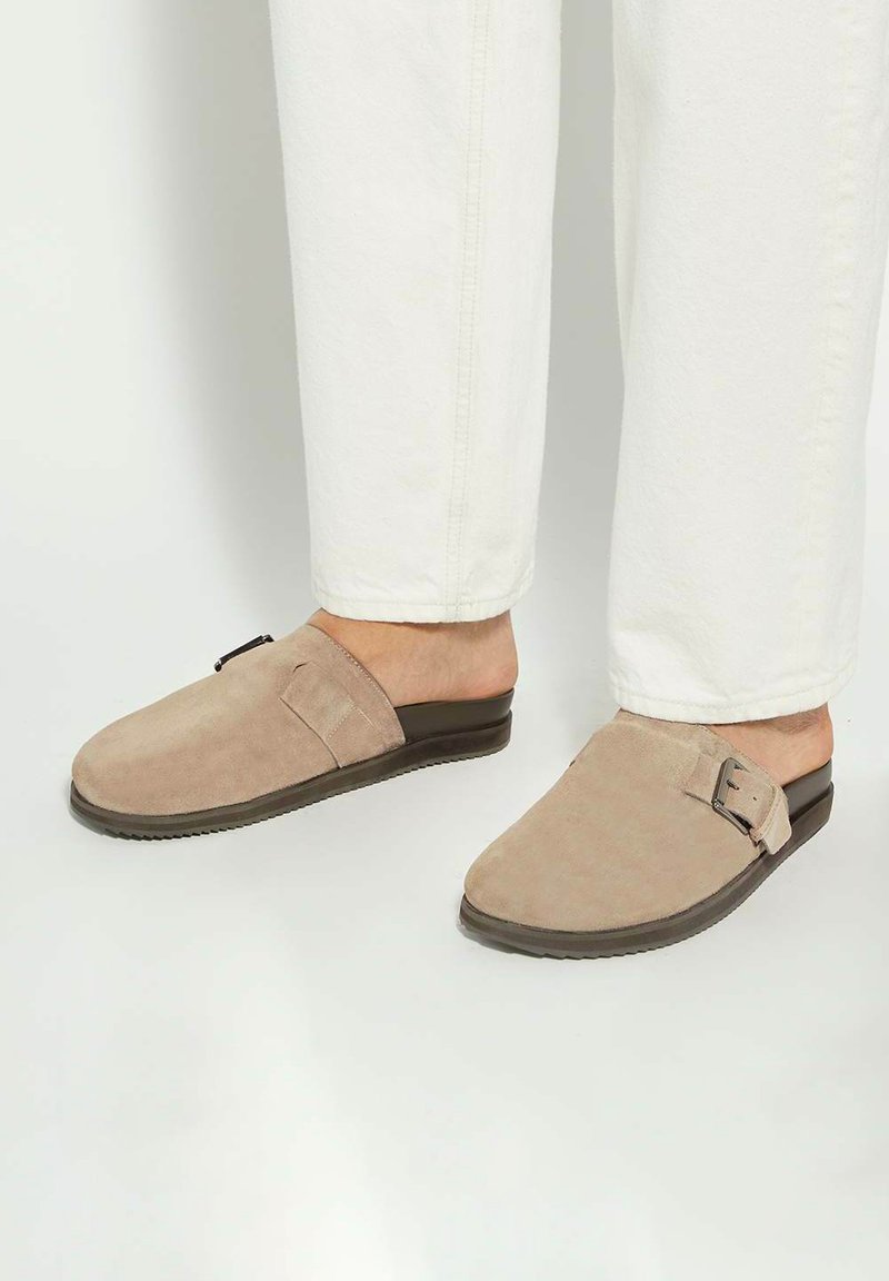 Beige suede slip-on shoes with a flat sole, featuring a side buckle detail and a smooth upper, paired with white pants.