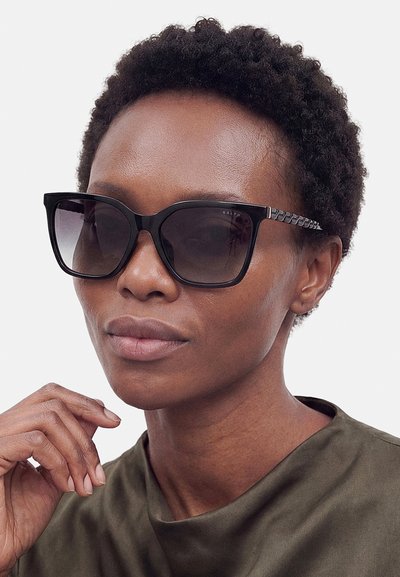 Vrouw met kort krullend haar die grote zwarte zonnebrillen en een olijfgroene top draagt, terwijl ze met haar hand haar kin aanraakt.