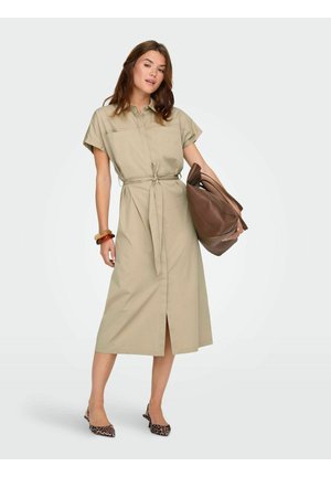 Nainen pukeutunut beigeen midipituiseen mekkoon, jossa on vyö, kantaa suurta ruskeaa nahkalaukkua ja käyttää rannekoruja ja leopardikuvioisia avokkaita.