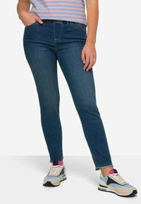 Jeans de mezclilla azul oscuro con un ajuste slim, que cuentan con un diseño clásico de cinco bolsillos y dobladillos deshilachados en los tobillos. Usados con zapatillas multicolores.