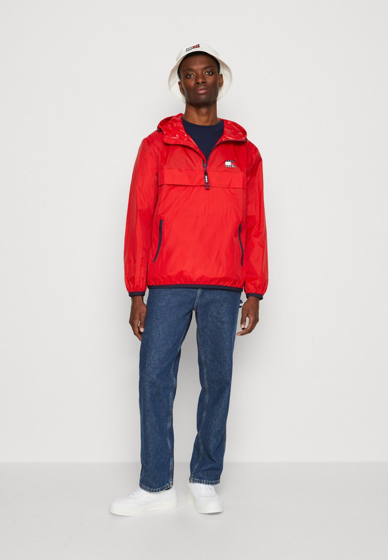Rød letvægts vindjakke med halv lynlås, forlommer og elastiske manchetter; parret med blå jeans og hvide sneakers. Bøllehat i hvid.