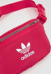 Rosa nylon midjeväska med en silverdragkedja, som har en vit Adidas-logga och tre vita randdesign. Justerbar rem ingår.
