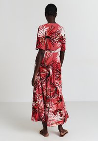 Robe maxi avec un fond rose et des motifs de feuilles tropicales rouges et blanches. Manches courtes et taille cintrée. Tissu léger.