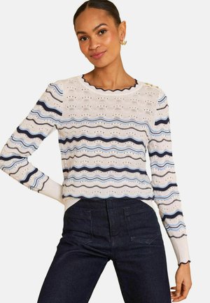 Vrouw draagt een crèmekleurige trui met marineblauwe en blauwe golvende strepen, gegolfde halslijn en manchetten, gecombineerd met donkere, hooggesneden jeans.