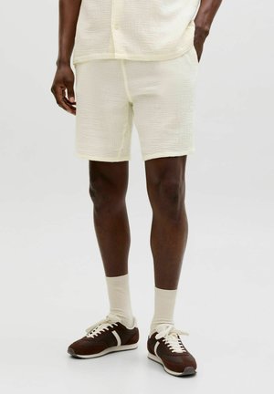 Hombre vistiendo pantalones cortos texturizados color blanco hueso y camisa a juego, combinados con calcetines blancos y zapatillas de deporte marrón oscuro con franjas y cordones blancos.