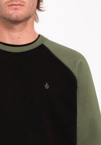 Sweatshirt noir avec des manches raglan vertes. Présente un tissu texturé et un petit logo sur la poitrine. Ce design offre une allure sportive.