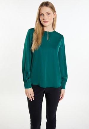 Femme aux longs cheveux blonds portant un chemisier vert à manches longues avec un décolleté en trou de serrure et un pantalon noir, se tenant devant un fond blanc.