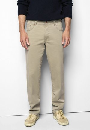 Mann trägt beige Hose, hellbeige Sneakers und einen dunklen Pullover mit langen Ärmeln und steht vor einem weißen Hintergrund.