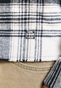 Das karierte Gewebe weist schwarze, weiße und orange Linien auf und enthält ein metallisches Akzentdetail. Der ausgefranste Saum fügt Textur hinzu. Beige Denim ist darunter sichtbar.