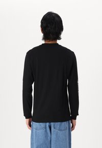 T-shirt noir à manches longues en coton, avec un col rond et un imprimé graphique sur la manche gauche. Pas de détail de texture visible.