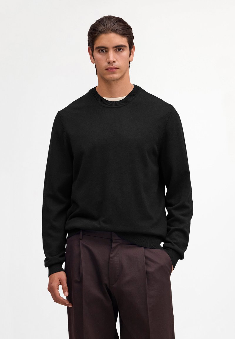 Ung mand med mørkt hår iført en sort rundhalset sweater og mørkebrune bukser, stående med den ene hånd i lommen foran en hvid baggrund.