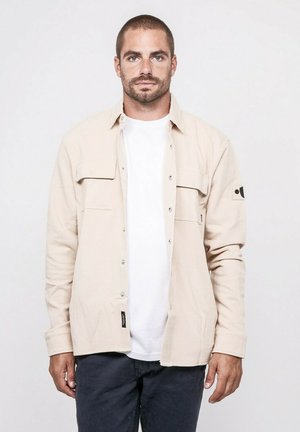 Homme aux cheveux courts et à la barbe, portant une chemise beige à boutons par-dessus un t-shirt blanc et un pantalon foncé, debout devant un fond uni.