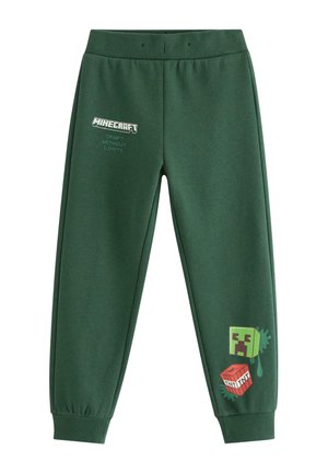 Junger Junge lächelt, während er einen grünen Minecraft-Sweatshirt und passende Hosen mit roten und grünen pixeligen Grafiken sowie braunen Sneakers trägt.