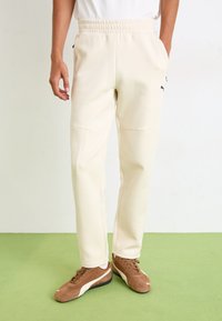 Cream-colored joggers with an elastic waistband, side pockets, and a black logo detail. The fabric appears soft and smooth.