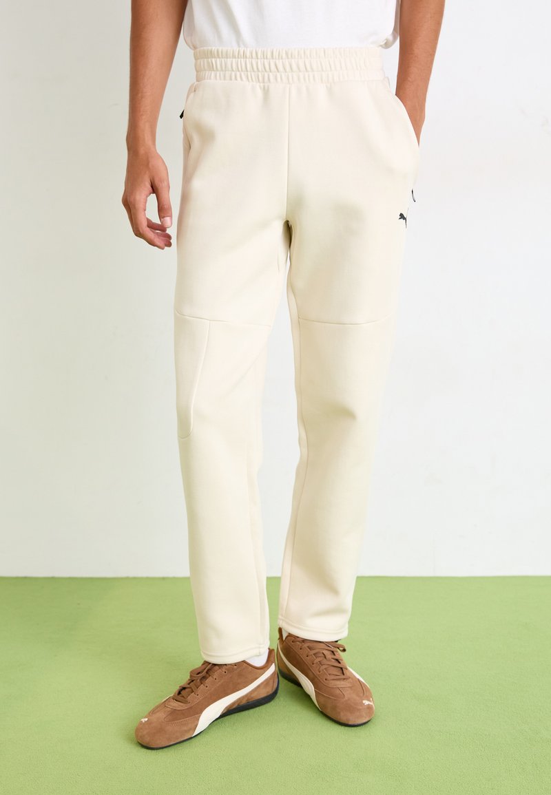 Cream-colored joggers with an elastic waistband, side pockets, and a black logo detail. The fabric appears soft and smooth.