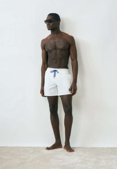 Άνδρας χωρίς πουκάμισο, φοράει λευκά μαγιό σορτς με μπλε κορδόνι και γυαλιά ηλίου, στέκεται ξυπόλητος μπροστά σε απλή ανοιχτόχρωμη τοίχο.