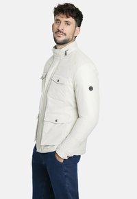 Chaqueta de plumas blanca con un collar alto, cuatro bolsillos frontales y un parche de logo redondo en la manga. Textura suave y silueta ajustada.