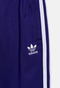 Fialové sportovní šortky ze lehkého materiálu s bílými bočními pruhy a vyšitým logem Adidas v bílé barvě na levé nohavici.