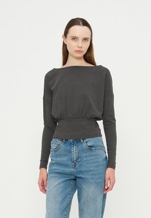 Jeune femme aux longs cheveux bruns portant un haut gris foncé à manches longues et un jean taille haute bleu clair sur un fond blanc uni.