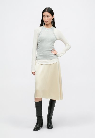 Top de manga larga gris con canalé, superpuesto a una falda midi de seda drapeada en color crema, combinado con botas negras hasta la rodilla. Diseño minimalista, texturas suaves.