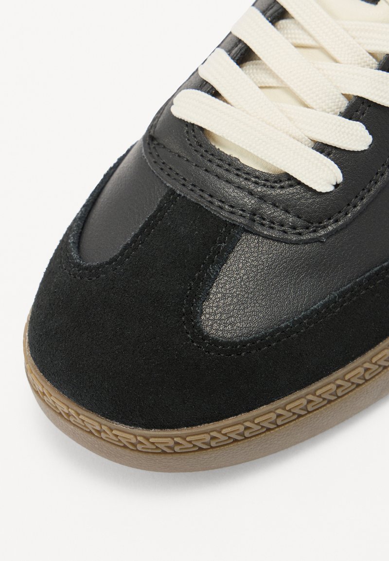 Zwarte sneaker met leren en suede materialen, crèmekleurige veters, gestructureerde gumrubberen zool en een gebogen ontwerp met gestikte accenten.