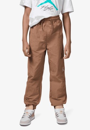 Barn iført brune bukser med snor, hvide og brune sneakers og en hvid t-shirt med en blå "Flight" grafik.