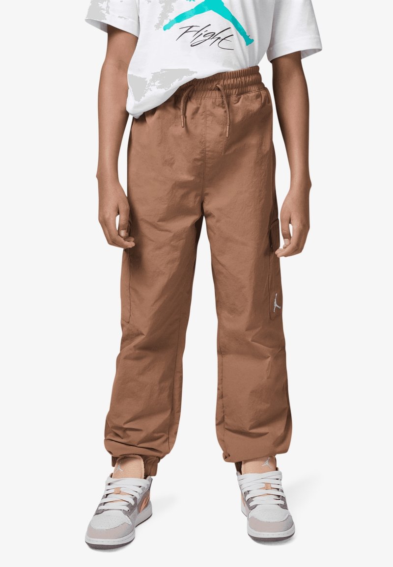 Enfant portant un pantalon cargo marron à cordon de serrage, des baskets blanches et marron, et un t-shirt blanc avec un motif bleu « Flight ».