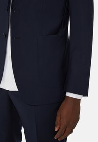 Isik, kellel on seljas tume mereväe sinine ülikond, valge särk, mis on nähtav nii manseti kui ka kere juures, käsi on lõdvasti reie kõrval.