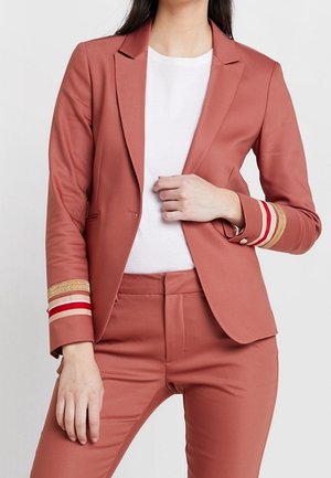 Blazer aderente color rosato con accenti a strisce dorate e rosse sulle maniche, abbinato a pantaloni su misura coordinati e una maglietta bianca. Texture liscia.
