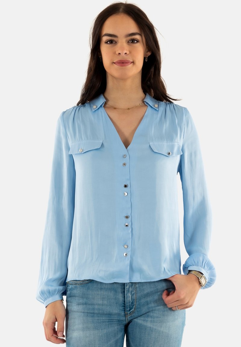 Blusa azzurra a maniche lunghe con scollo a V, due tasche sul petto, bottoni in argento sulla parte frontale e polsini arricciati. Texture satinata.