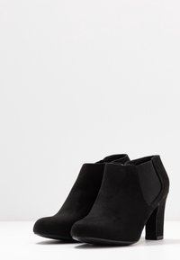 Černé semišové kotníkové boty s kulatou špičkou, elastickými bočními panely a blokovým podpatkem o výšce 8,9 cm. Hladký povrch a minimalistický design.