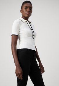 White ribbed polo shirt with a black collar and button placket, featuring short sleeves and black trim, paired with black trousers.