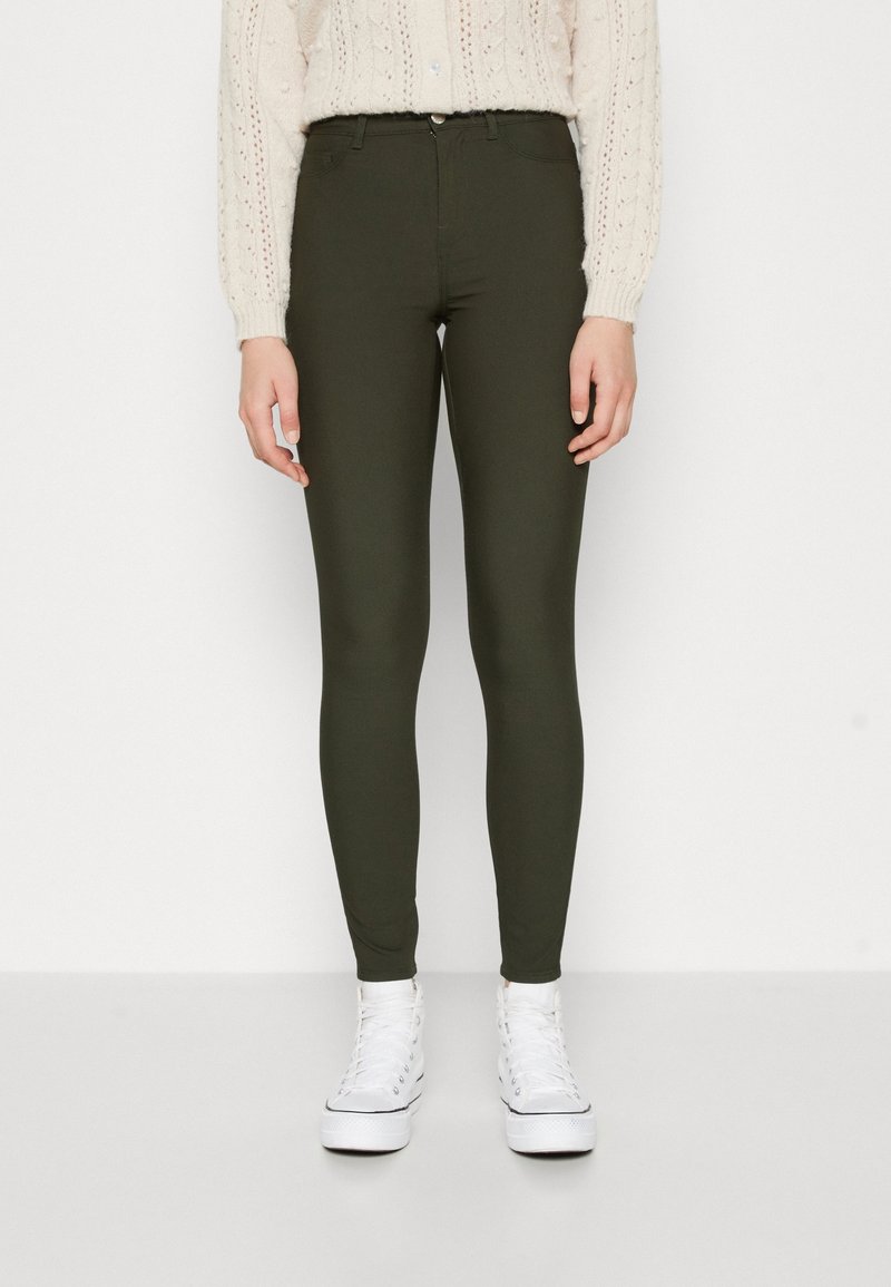 Jean skinny vert olive en tissu extensible, avec une taille haute, des poches avant et une texture lisse, associés à des baskets blanches.