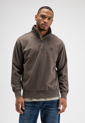 Mann trägt braunen Viertel-Reißverschluss-Pullover über einem beigen Hemd und blauen Jeans und steht vor einem schlichten hellen Hintergrund.