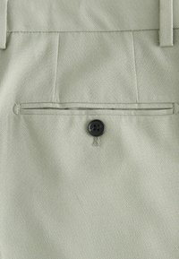 Gros plan d'un pantalon en tissu gris clair montrant une poche passepoilée arrière avec un bouton noir et des passants de ceinture au-dessus.