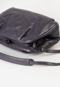Lila Lederhandtasche mit glatter Textur, Reißverschluss und verstellbarem Tragegurt. Mit dezentem Branding, das auf dem Gurt geprägt ist.