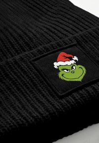 Schwarze Strickmütze mit einem rechteckigen Patch, der eine grüne Grinch-Figur mit rotem Weihnachtsmannhut zeigt, mit strukturiertem Stoff und filigranen Details.
