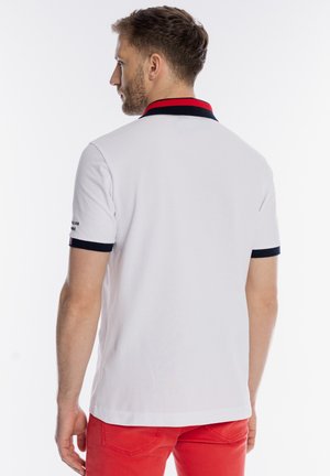 Mężczyzna zwrócony tyłem, ubrany w białą koszulkę polo z czarnym i czerwonym kołnierzykiem oraz czerwone spodnie na białym tle.