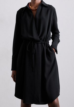 Czarna długa sukienka kopertowa z długim rękawem, kołnierzykiem i paskiem w talii, noszona z masywnym czerwono-złotym naszyjnikiem z koralików.