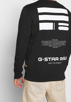 Mężczyzna ubrany w czarną bluzę G-Star Raw z białym geometrycznym wzorem i napisem na plecach, do tego jasnobeżowe spodnie.