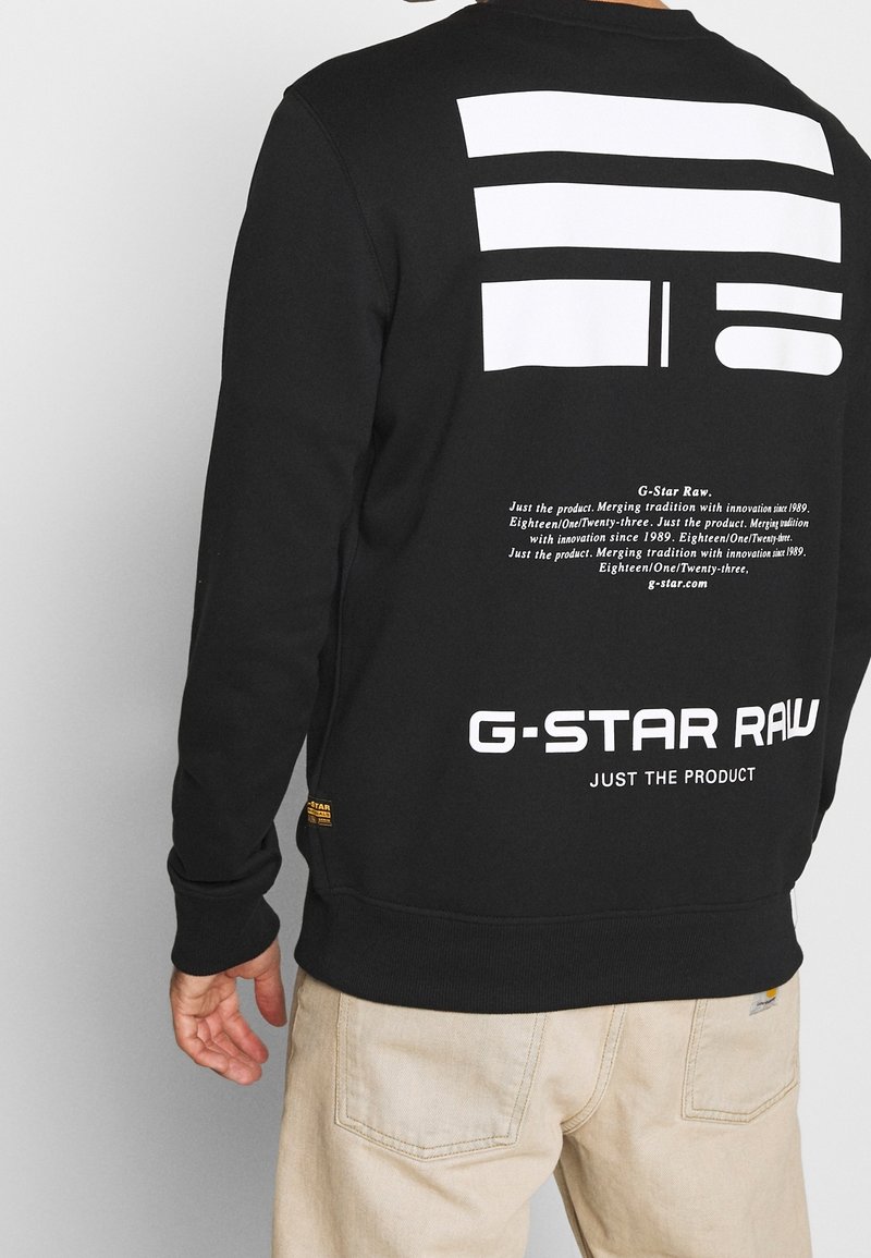 Homme portant un sweat-shirt noir G-Star Raw avec un motif géométrique blanc et un texte au dos, associé à un pantalon beige clair.