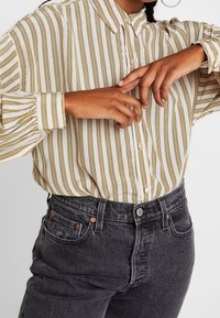 Camicia con bottoni a righe verticali gialle e marroni, maniche lunghe e colletto classico, abbinata a jeans in denim scuri a vita alta.