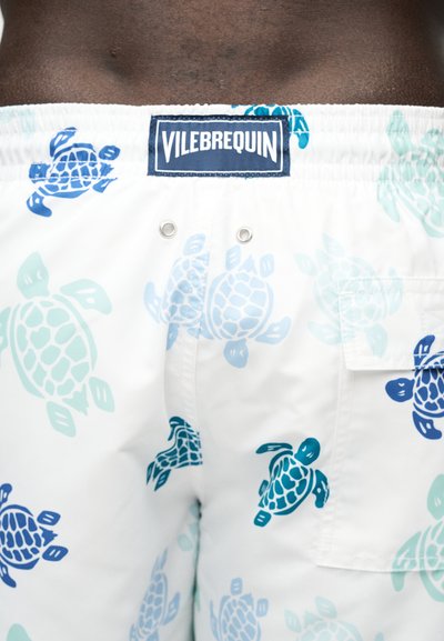 Λευκά μαγιό σορτς με σχέδια μπλε και πράσινης θαλάσσιας χελώνας και ετικέτα της μάρκας Vilebrequin στη μέση.