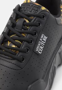 Baskets noires en cuir perforé avec une semelle en caoutchouc. Elles disposent de lacets noirs, d'accents texturés et d'un intérieur à motifs avec des détails jaunes.