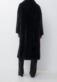 Cappotto lungo nero realizzato in materiale di pelliccia sintetica dalla texture, con ampio colletto e spalle scese, abbinato a pantaloni neri aderenti.